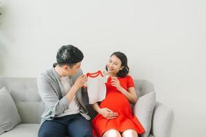 Portrait of a happy pregnant woman holding baby t-shirt and of her husband on a sofa at home photo