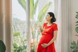 Young Asian pregnant holding phone and standing behind the window. photo