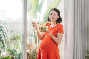 lleno de vitaminas. mujer embarazada enérgica y guapa comiendo su carne mientras lleva un plato en una mano y se relaja contra la ventana foto