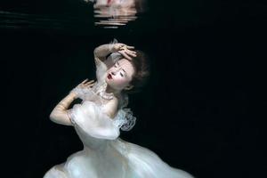 mujer joven en vestido largo de lujo blanco bajo el agua en el fondo negro. movimiento, arte, concepto de invierno foto