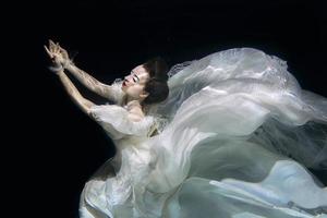 young woman in long white luxury dress underwater on the black background. Motion, art, winter concept photo