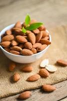 Delicious sweet almonds on the wooden table, roasted almond nut for healthy food and snack, Almonds nuts on white bowl and green leaf on sack background photo