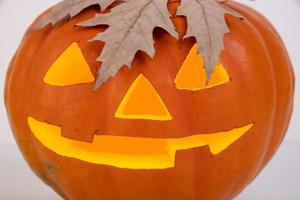 calabaza naranja con ojos luminosos, nariz y boca y hoja de arce otoñal en forma de cabello sobre un fondo blanco. Víspera de Todos los Santos foto