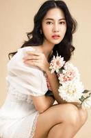 mujer asiática con una cara hermosa y una piel limpia y fresca perfecta con flores. lindo modelo femenino con maquillaje natural y ojos brillantes sobre fondo beige aislado. foto