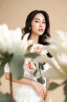 mujer asiática con una cara hermosa y una piel limpia y fresca perfecta con flores. lindo modelo femenino con maquillaje natural y ojos brillantes sobre fondo beige aislado.
