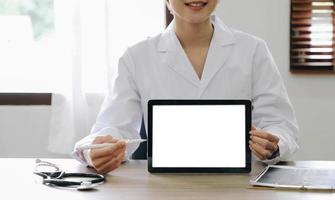 retrato de una doctora experimentada, cardióloga, presenta una tableta de pantalla blanca de tecnología moderna que usa para consultar a los pacientes, que usan un estetoscopio uniforme de bata blanca en la sala de examen. foto