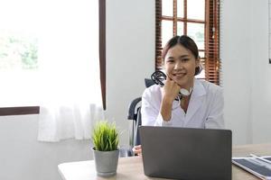 Smiling asian female doctor with laptop computer in her office. Friendly medical professional with tablet computer in clinic. photo