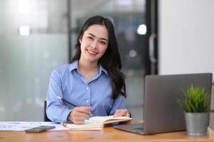 smiling beautiful young Asian businesswoman sitting with laptop and computer while doing some paperwork at the office. Looking at camera. photo