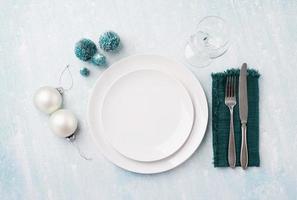 Christmas table setting with white dishware, silverware and green decorations on blue background. Top view. photo