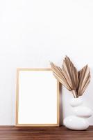 White frame mockup with dry palm leaves in a vase on wooden table photo