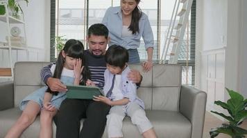 Happy Asian Thai family, parents, and children have fun using digital tablet together on sofa in home living room, a lovely leisure weekend, and domestic wellbeing lifestyle with internet technology. video