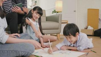 Asian Thai siblings and mum are sitting on living room floor, drawing with colored pencils together, dad leisurely relaxing on a sofa, lovely happy weekend activity, and domestic wellbeing lifestyle. video