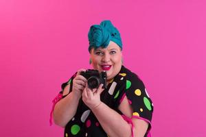 mujer sonriente de talla grande con expresión divertida de la cara emocional con cámara vintage en vestido de mezclilla aislado en fondo rosa, viajero de vacaciones, estilo de moda de verano, turista emocionado. foto