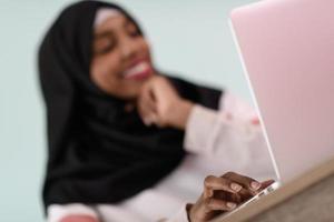 afro muslim  woman  wearing a hijab sits smiling in her home office and uses a laptop photo
