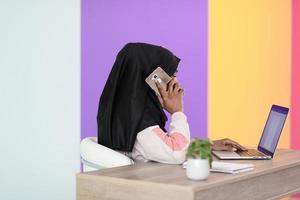 afro muslim  woman  wearing a hijab sits smiling in her home office and talking on smartphone photo