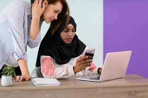 una chica afro con hiyab y una mujer europea usan un teléfono celular y una computadora portátil en la oficina de su casa foto