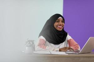 afro muslim  woman  wearing a hijab sits smiling in her home office and uses a laptop photo