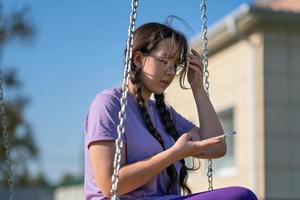 a teenager girl swings on a swing with a phone in her hands, communicates in social networks or learns online photo