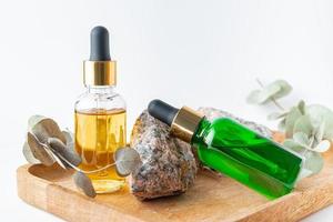 glass dropper bottles with a pippette with gold rubber tip and green eucalyptus leaves near it on wooden tray on white background. Nature Skin concept. Organic Spa Cosmetics. photo