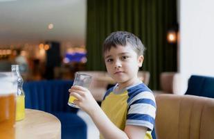 Portrait Young kid drinking fresh orange juice for breakfast in cafe, Happy child boy holding glass of fruit juice while waiting for food in restaurant, Healthy food lifestyle concept photo