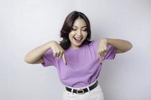 Excited Asian woman wearing lilac purple t-shirt pointing at the copy space downside her, isolated by white background photo