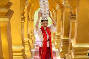 joven asiática con traje tradicional birmano sosteniendo un tazón de arroz a mano en la pagoda dorada en el templo de myanmar. mujeres de myanmar sosteniendo flores con vestido tradicional birmano visitando un templo budista foto