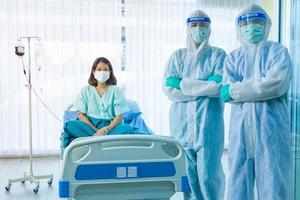 Asian woman patient feel happy because get well and recovered from COVID-19 or CORONA virus diease. Two medical staffs wear PPE suit standing beside patient bed and looking at the camera photo