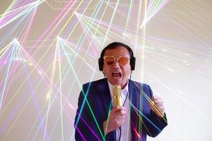 Portrait of the handsome attractive singer in a suit and brown glasses, singing hit with open mouth in gold microphone gesture with hand isolated on grey background and shine color light. photo