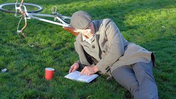 jovem hipster masculino com bicicleta retrô ler livro no parque outono video