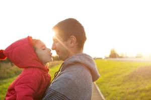 Little girl in red jacket with a hood hugs and kisses her dad, smiles, touches her nose. Happy family, children's emotions, father's day, bright rays of the sun, Caucasian appearance. Space for text.