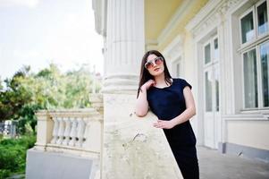 Brunette girl at black dress, sunglasses posed against old vintage house, at street of city. photo