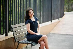 Brunette girl at black dress sitting on bench and posing at street of city. photo