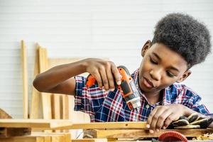 retrato de un niño afroamericano que usa camisa con un taladro en las manos y ayuda a papá a armar un estante de muebles con una herramienta de destornillador eléctrico, concepto de aprendizaje. foto