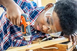 Portrait of african american little child wearing shirt standing with a drill in hands and help dad assembling furniture shelf with power screwdriver tool, learning concept. photo