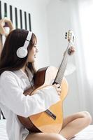 retrato joven sonrisa feliz hermosa mujer asiática relajarse dormitorio. mujer con auriculares tocando la guitarra acústica en el dormitorio blanco foto
