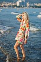 Blonde woman wearing dress standing on sand beach at sea shore enjoying view of sunset. photo