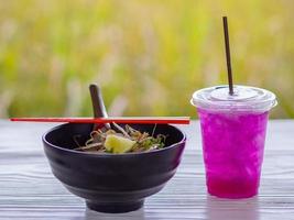 Noodle soup in black cup with red chopsticks and herb water on white wooden table. photo