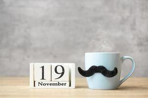 día internacional del hombre con calendario del 19 de noviembre, taza de café azul o taza de té y decoración de bigote negro en la mesa. feliz día del padre y concepto de celebración foto