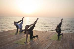 group of womans and man doing yoga on wooden pier, morning sea background photo