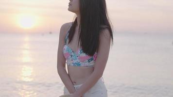 Close up asian cute bikini woman slowly walking against the golden sunset beach, a solo female tourist enjoying watching sunset before leaving the peaceful seashore at evening time, holiday isolation video