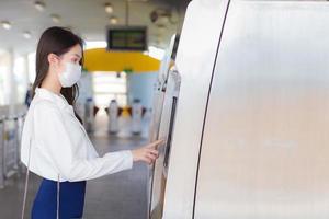 Beautiful young Asian businesswoman in a white shirt stands wear a mask to prevent disease virus and dust pm.2.5 to press a ticket vending machine in a sky train station to go to work at the office. photo