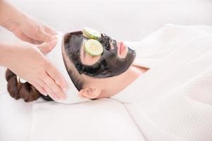 una hermosa mujer asiática usa barro de spa y pepino para el tratamiento facial foto