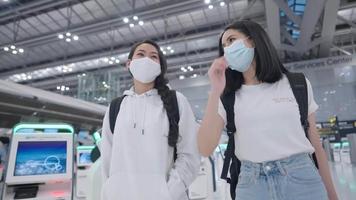 Asian friends wear facemask walking together inside airport terminal, travel insurance package, backpack travellers, new normal way for travel after pandemic crisis, infections disease risk prevention