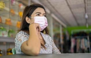 una mujer tailandesa con una máscara rosa se sienta cerca de una mesa esperando clientes. foto