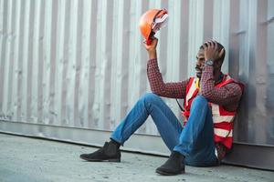 técnico o ingeniero afroamericano. siéntese cerca de un contenedor y luzca cansado y somnoliento o desempleado. concepto de transportador de carga industrial de logística. foto