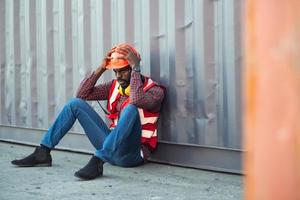 técnico o ingeniero afroamericano. siéntese cerca de un contenedor y luzca cansado y somnoliento o desempleado. concepto de transportador de carga industrial de logística. foto