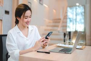 sonriente joven mujer de negocios asiática y empresaria usando teléfono móvil y desplazándose y navegando a través de las redes sociales tomando un descanso después de trabajar en una computadora portátil foto