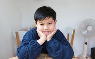 Cute Asian boy with white skin smiling looking in camera happily resting chins on hands-on wooden table. Close-up portrait of a cute child photo