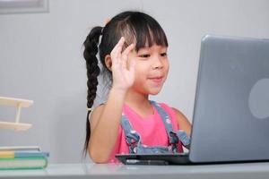 Smiling little Asian girl studying online having video call distant class with teacher using laptop. Happy little girl wave greeting with tutor while studying online with laptop at home. homeschooling photo