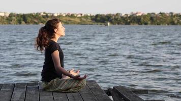 Seitenansicht einer schlanken jungen Frau, die im Lotussitz auf einem Pier sitzt und ihre Hände hebt. Frau, die Yoga am Strand bei Sonnenuntergang gegen das Wasser praktiziert. 4k-Zeitlupenvideo. video